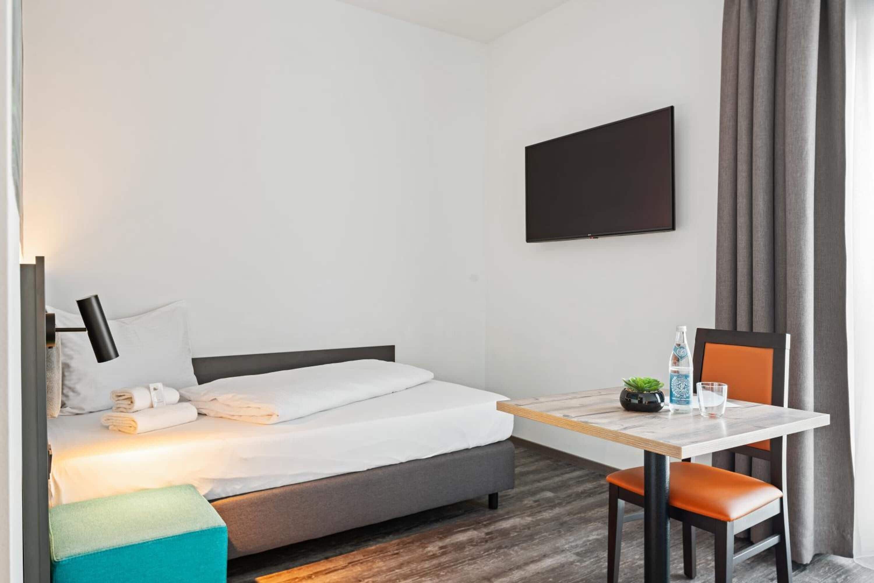 Habitación de hotel con cama, escritorio, silla, televisor y ventana con cortinas.