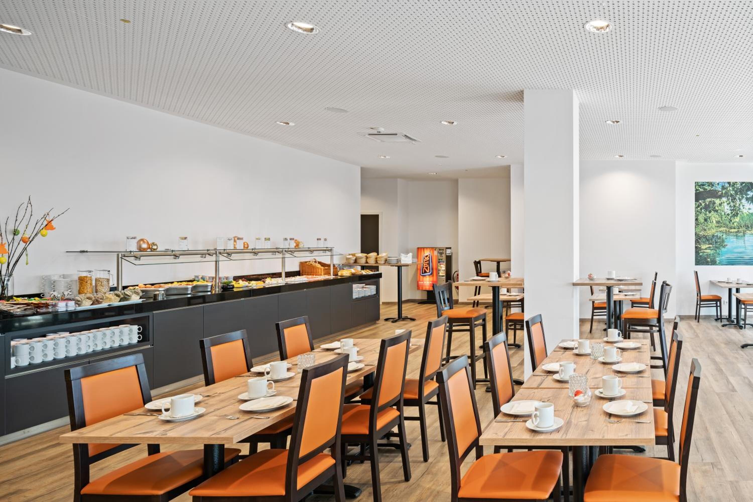A modern dining area with multiple tables and chairs, a buffet counter with various food items, and a large window with a view of greenery.