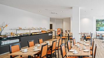 A modern dining area with multiple tables and chairs, a buffet counter with various food items, and a large window with a view of greenery.