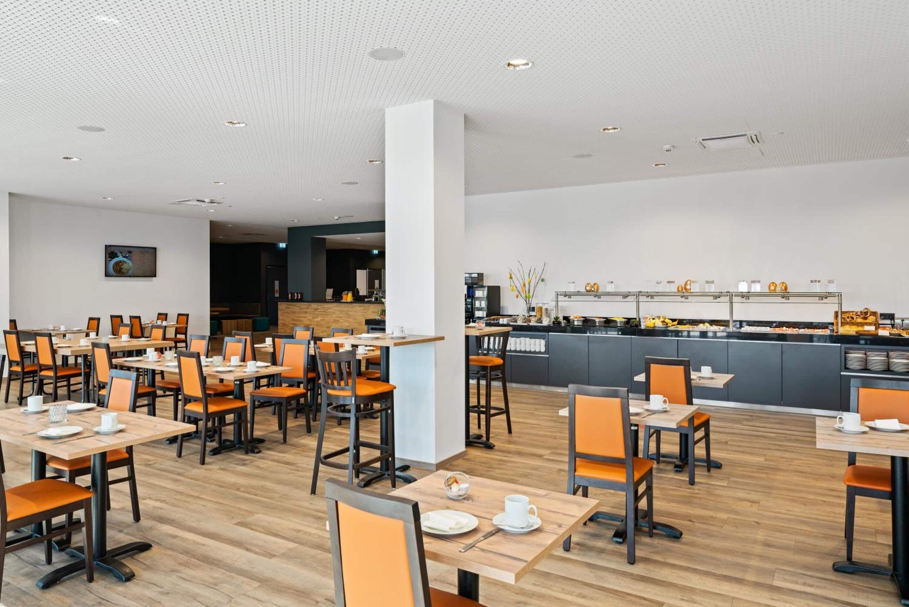 Zona de menjador d'un hotel modern amb diverses taules i cadires, una barra central amb racó de cafè, i una cuina al fons.