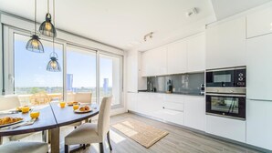 Eine moderne Küche mit weißen Schränken, ein Essbereich mit Tisch und Stühlen sowie ein Blick auf die Stadt durch große Fenster.
