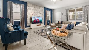 Una sala de estar moderna con un sillón azul, un sofá blanco, una mesa de centro de vidrio y un televisor de pantalla plana sobre un mueble blanco.