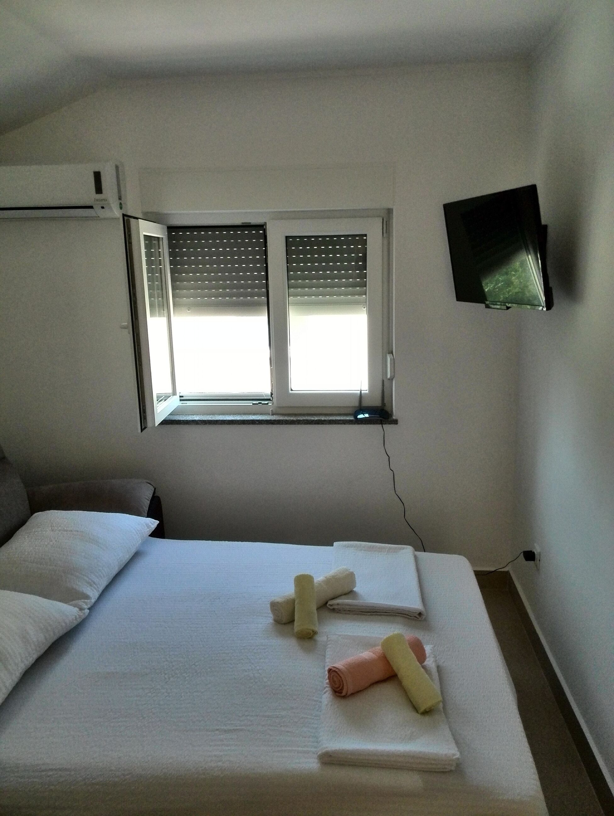 A hotel room with a bed, pillows, a window with blinds, a flat-screen TV mounted on the wall, and a power outlet on the wall.
