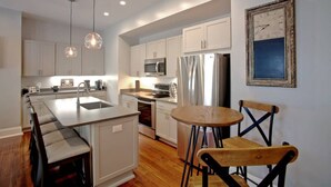 Una cocina moderna con armarios blancos, electrodomésticos de acero inoxidable, iluminación colgante y una zona de comedor con una mesa redonda y sillas de madera.