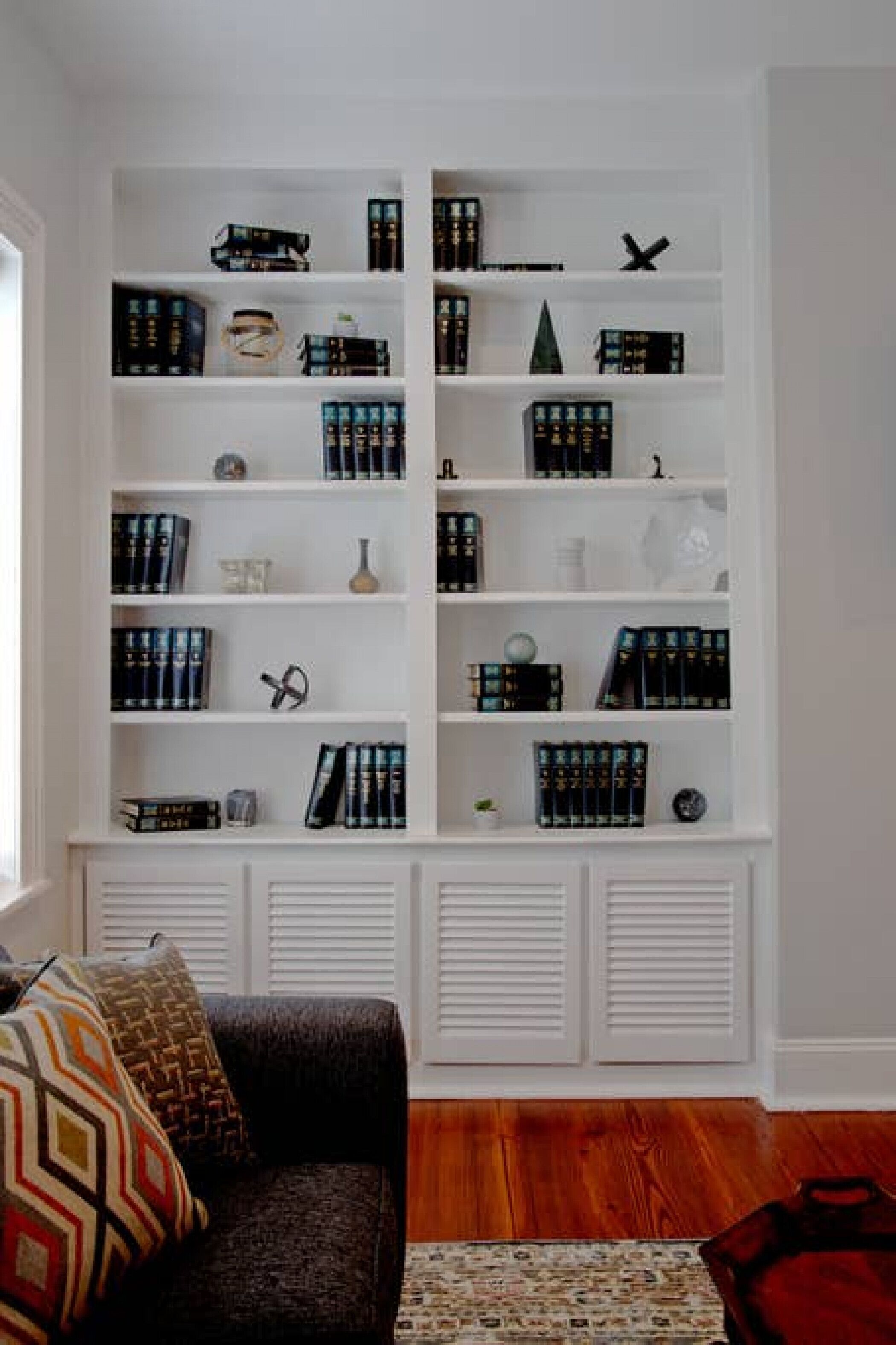 A room with a white built-in bookshelf filled with books and decorative items, a patterned sofa, and a wooden floor.