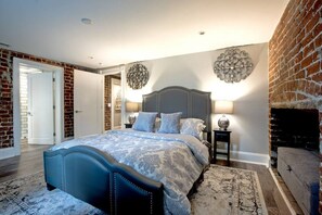 Une chambre avec un grand lit, un mur en briques, une cheminée et une porte donnant sur une autre pièce.