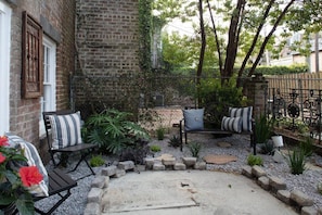 A brick building with a small patio area featuring two black metal chairs with striped cushions, a stone pathway, and a metal fence.
