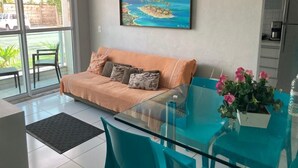 Un salon moderne avec une table à manger en verre, un canapé avec des coussins, un tableau représentant un paysage tropical et un coin cuisine avec un réfrigérateur.