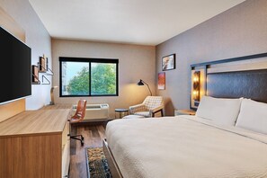 A hotel room with a bed, desk, chair, and a large window with a view of trees.