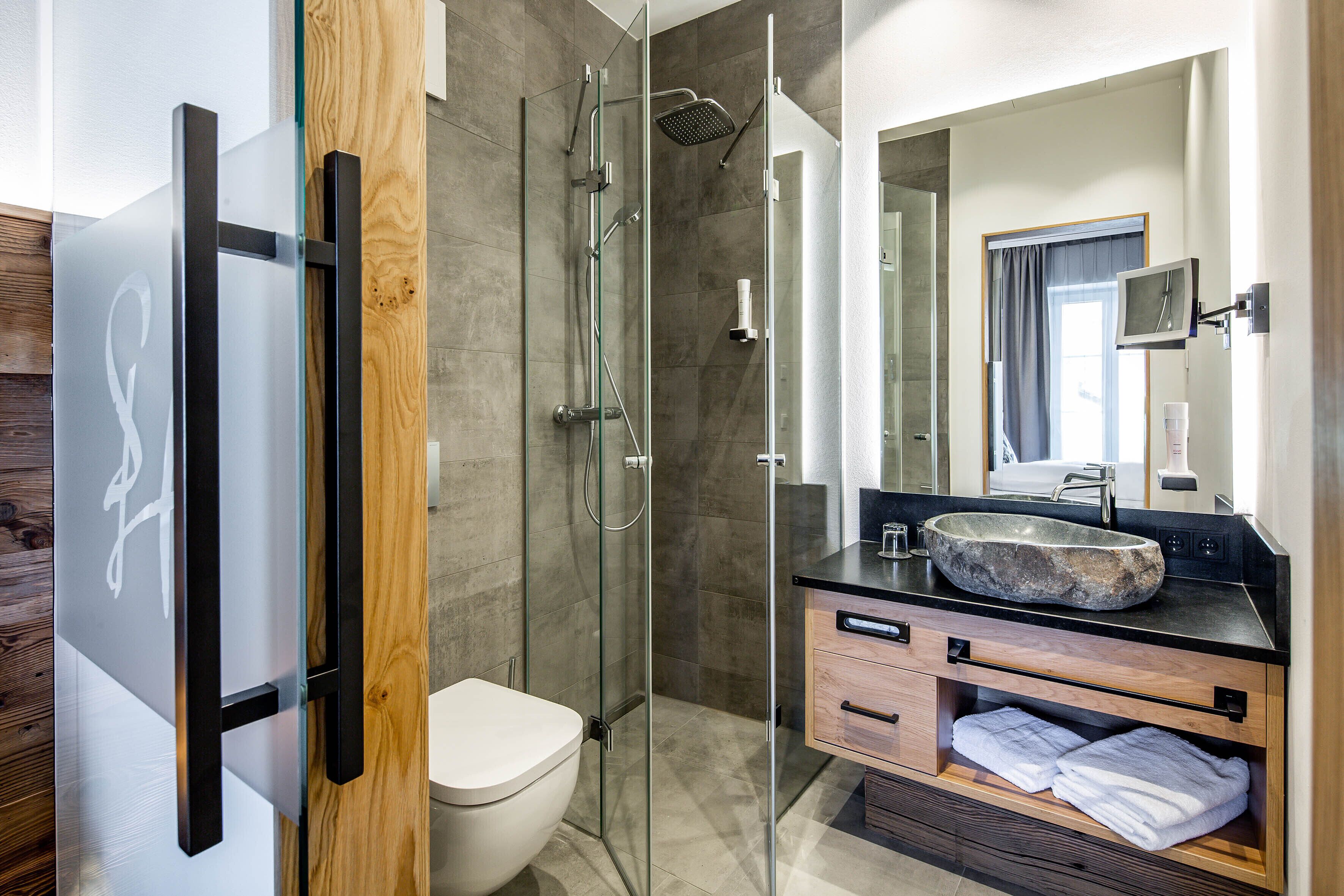 A modern bathroom with a glass shower enclosure, a white toilet, a large mirror, and a wooden vanity with a stone sink.