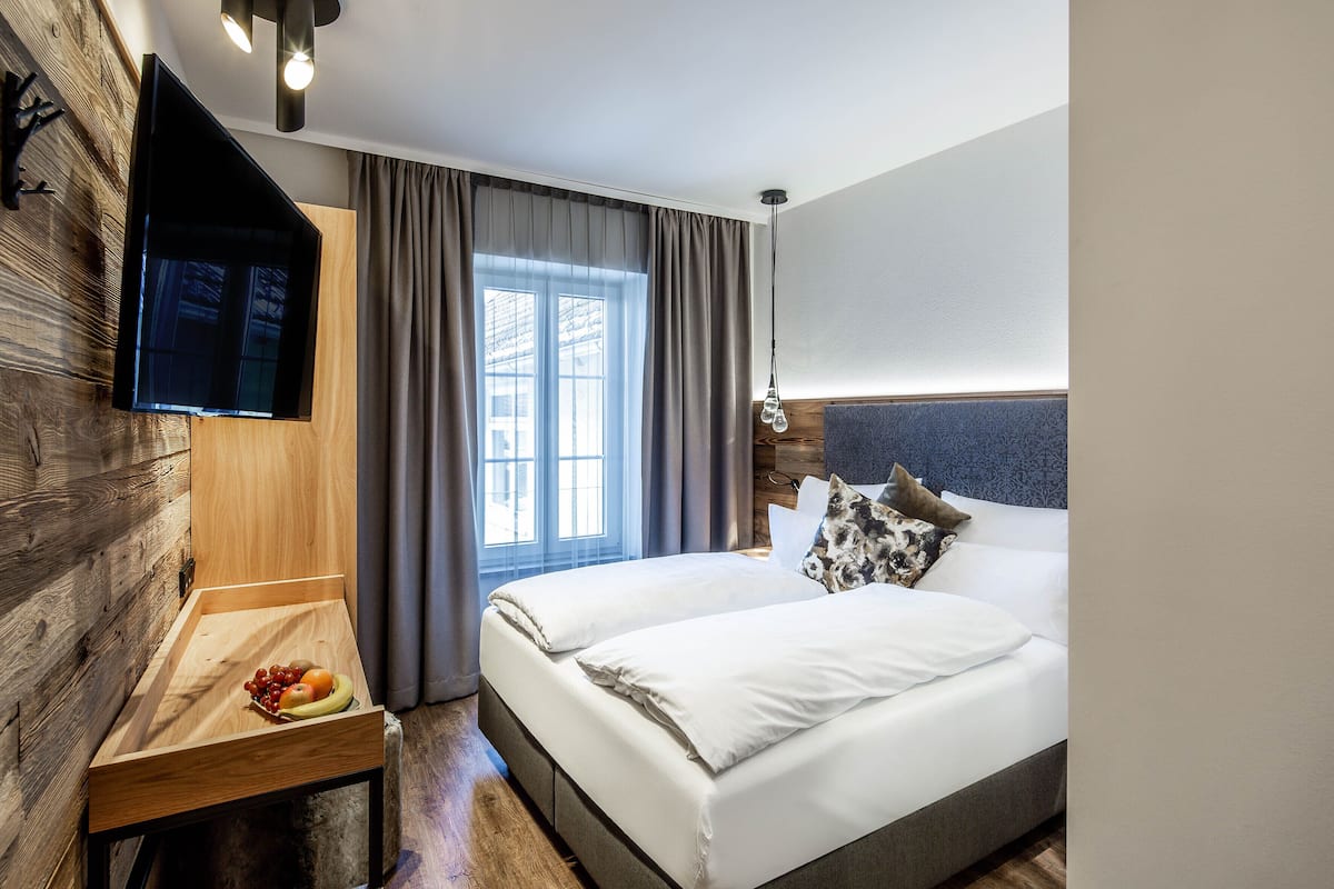 Une chambre d’hôtel moderne avec un mur d’accent en bois, un téléviseur à écran plat, une table de chevet avec des fruits, un grand lit avec une literie blanche et une fenêtre avec des rideaux.
