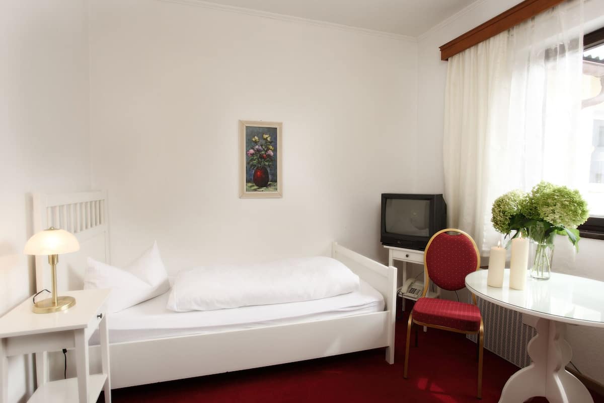 Une chambre d’hôtel avec un lit simple, un bureau avec une télévision, une chaise, une petite table ornée d’un vase de fleurs et un tableau accroché au mur.