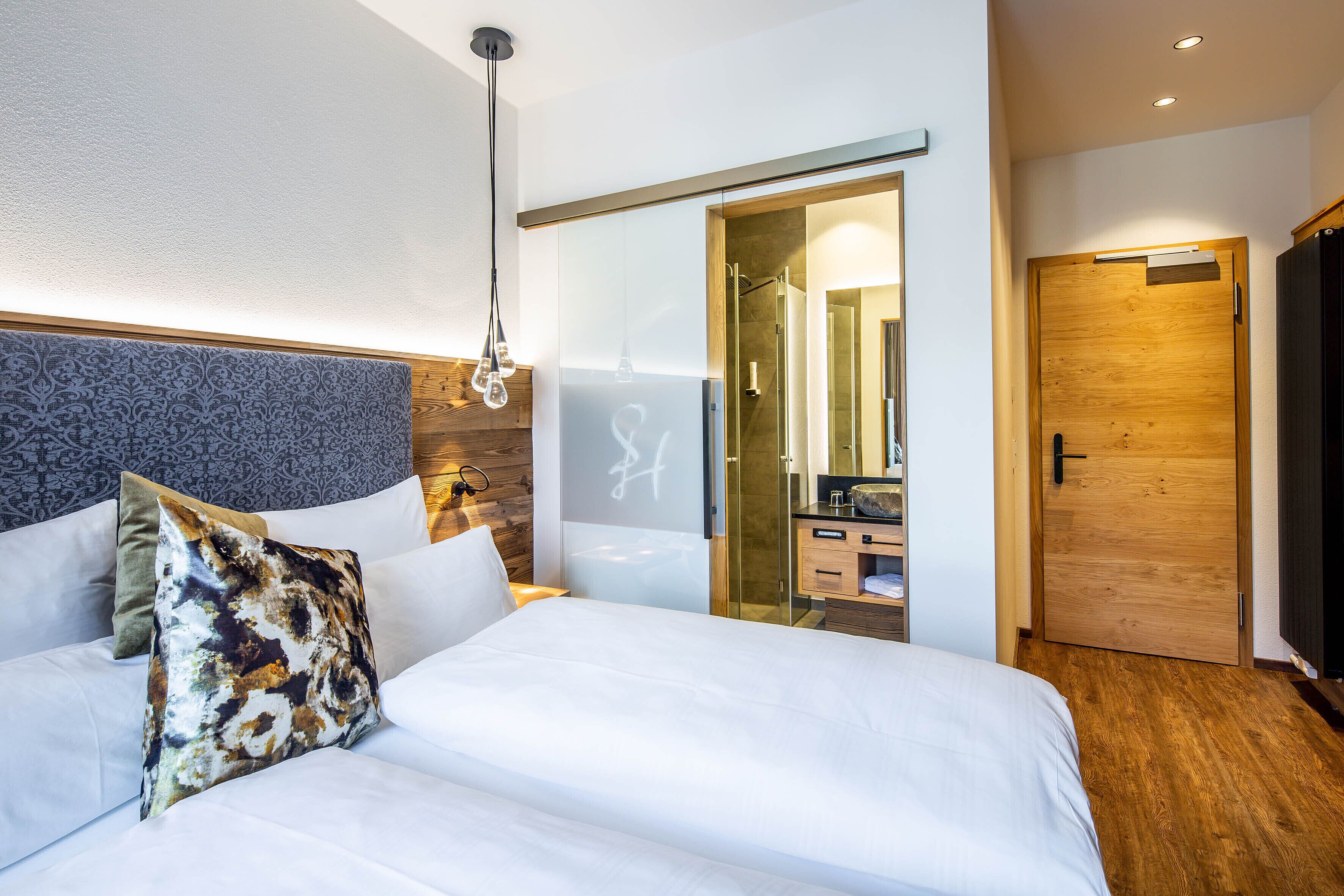 A modern hotel room with a large bed, a wooden headboard, a glass shower enclosure, and a wooden door.