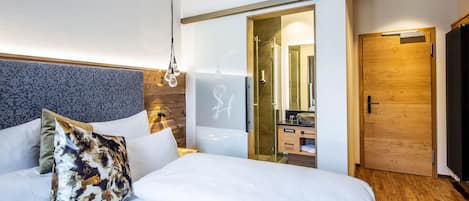 A modern hotel room with a large bed, a wooden headboard, a glass shower enclosure, and a wooden door.