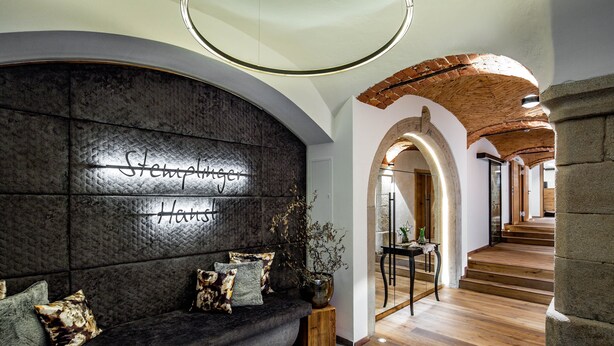 A modern interior with a dark textured sofa, a wall-mounted sign reading "Stempelberger Haus," and a wooden staircase leading to another level. - Stemplinger Hansl (Hauzenberg)