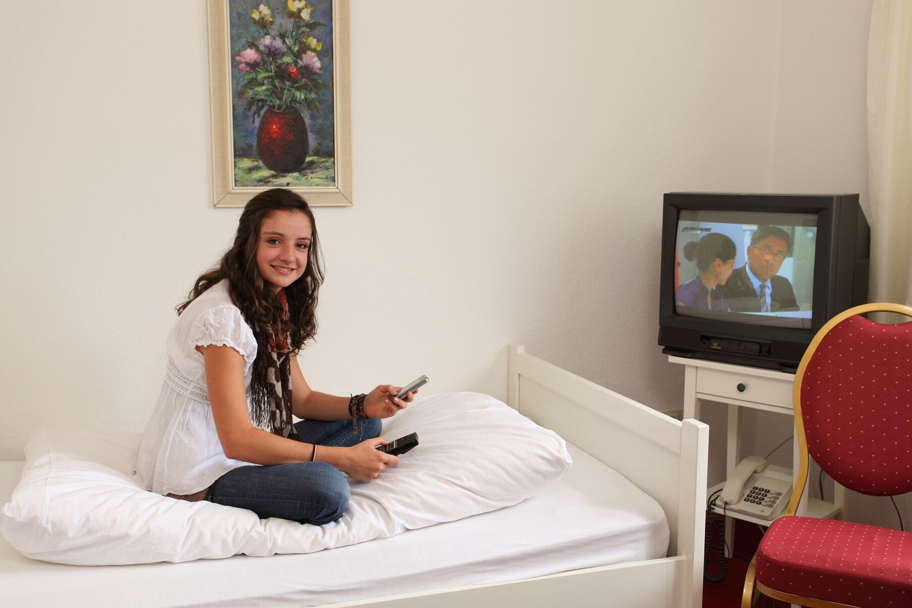 A person sitting on a bed in a room with a television, a telephone, and a chair.