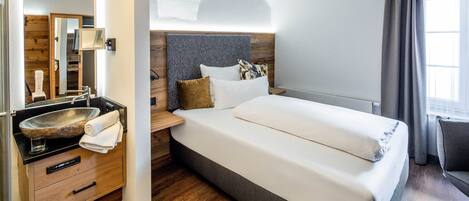 A modern hotel room with a large bed, a wooden vanity, a built-in sink, and a view of the outdoors.