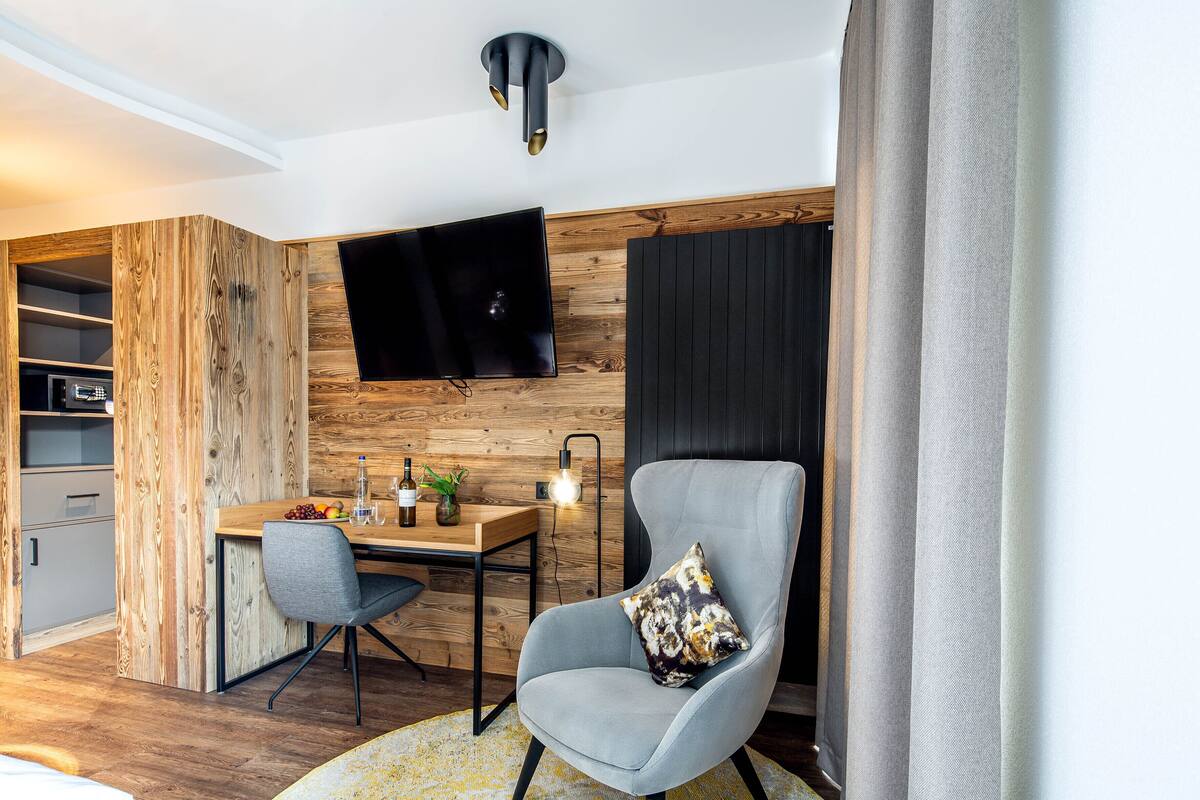 Un salon moderne avec un mur d’accent en bois, un téléviseur à écran plat fixé au-dessus d’un bureau en bois, un fauteuil gris agrémenté d’un coussin à motifs, et une petite table d’appoint avec une plante et une bouteille.