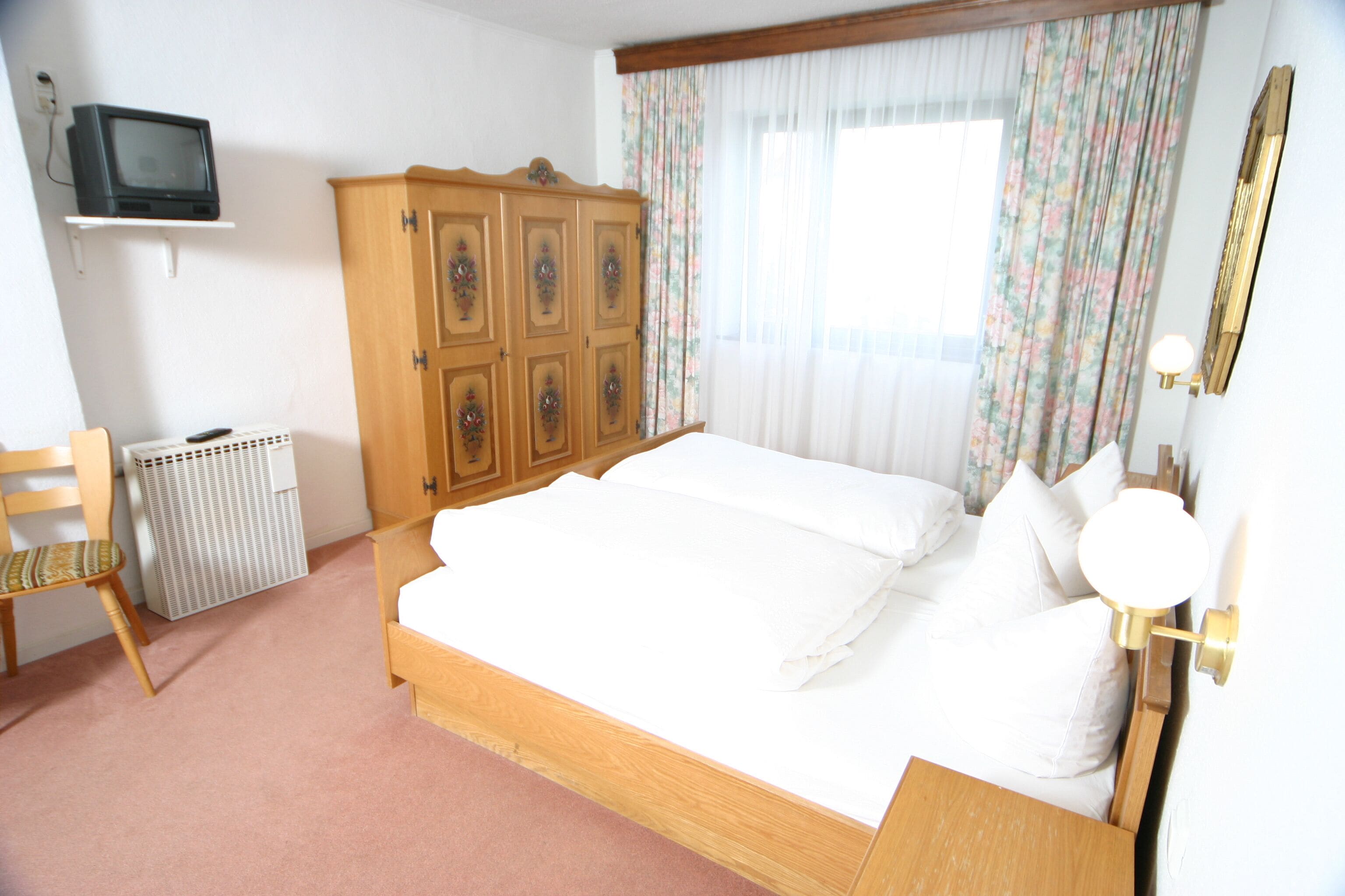Un dormitorio con cama de madera, una silla, un radiador, televisor sobre una estantería y una ventana con cortinas.