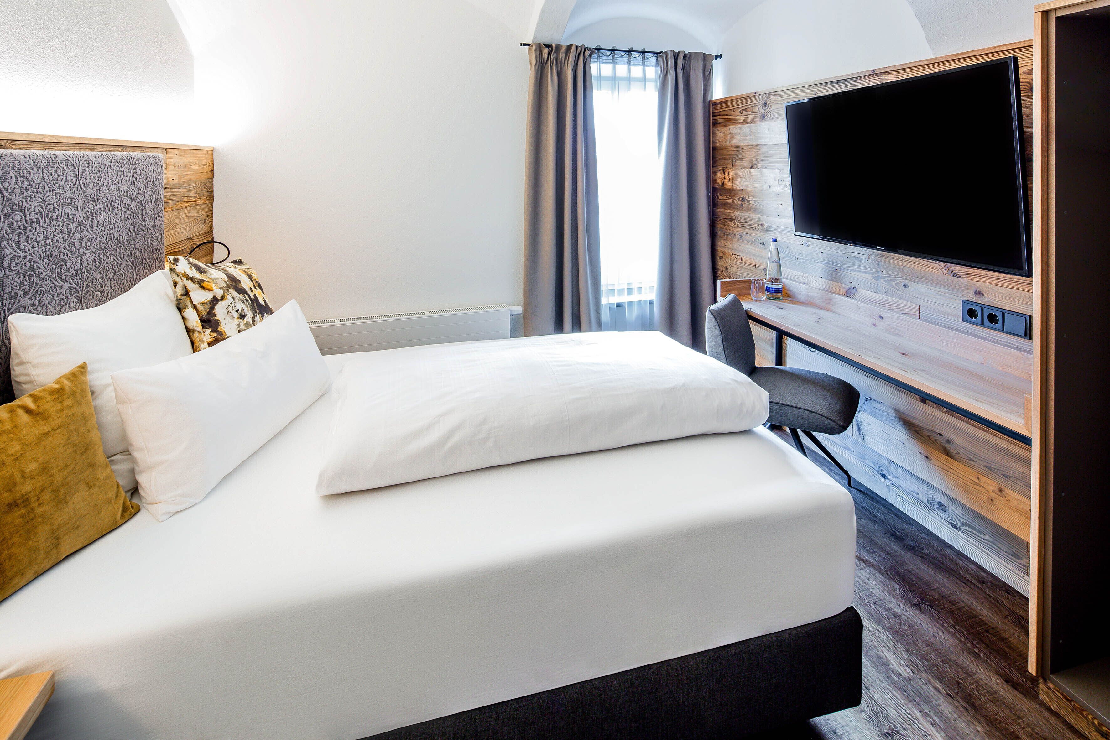 Ett hotellrum med en stor säng, en platt-TV som är monterad på en väggpanel i trä och ett fönster med gardiner.