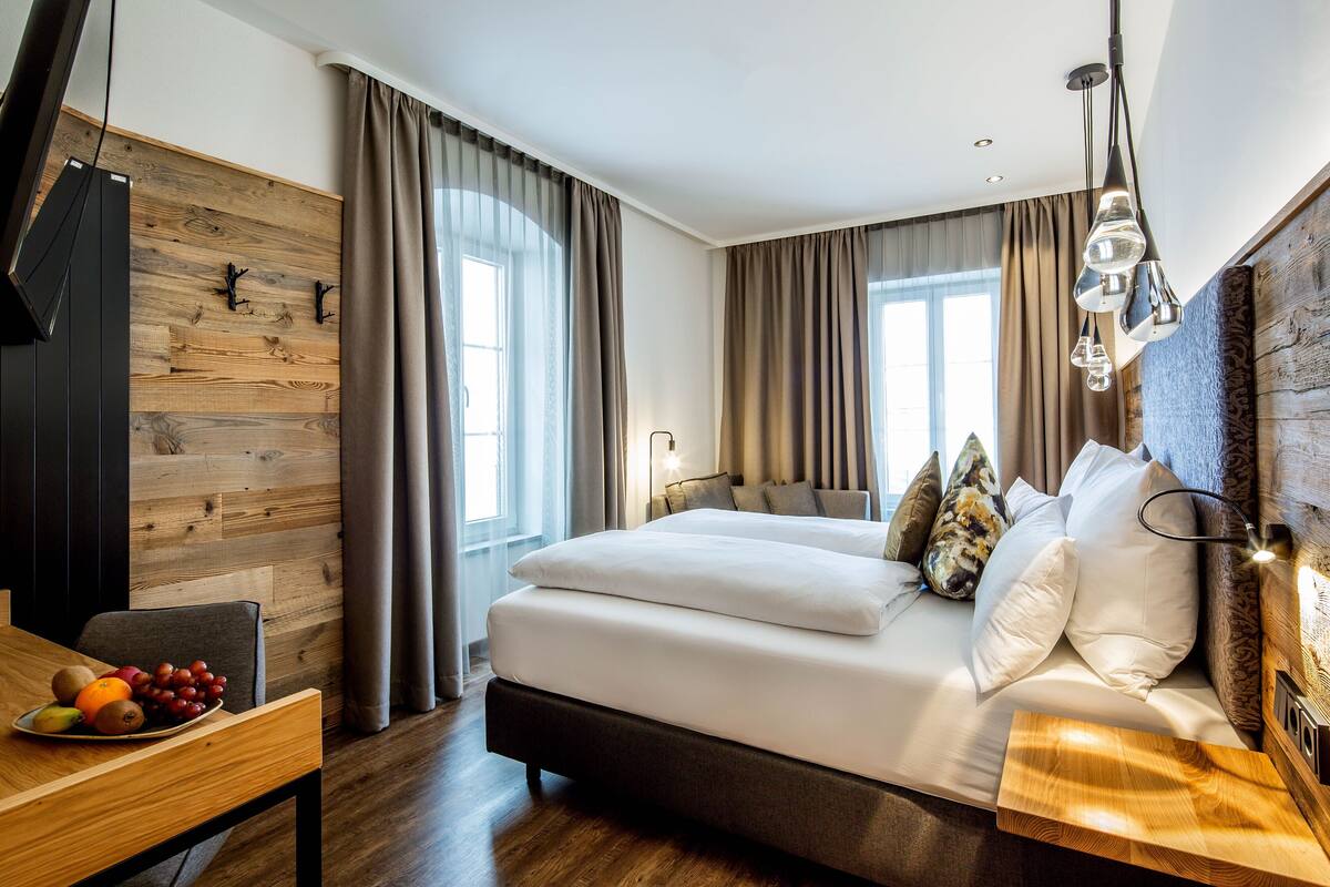 Une chambre d’hôtel moderne avec un mur d’accent en bois, un grand lit avec une literie blanche, un bureau en bois agrémenté d’un panier de fruits et un téléviseur placé à gauche.