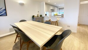 A modern dining area with a wooden table and black chairs, a kitchen in the background, and a white wall with a framed picture. - NEW FOR 2020 - 3 Harvest Moon is a stunning fully refurbished 2 bedroom, 2 en suite bathroom apartme (Newquay)
