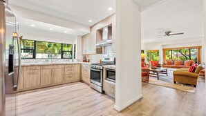 A modern kitchen with wooden cabinets, stainless steel appliances, and a large island. There is a dining area with a sofa and chairs, and a ceiling fan. - Spacious Poipu Beach Home near Hyatt-Remodeled Kitchen & Bathrooms Large Hot Tub (Koloa)