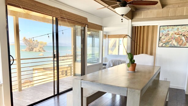 A room with a large sliding glass door leading to a balcony with a view of the sea. There is a table with a potted plant on it, and a ceiling fan above.