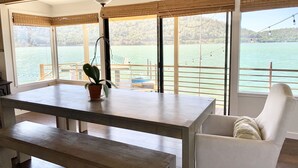 A room with a wooden table, a bench, a white chair, a ceiling fan, and large windows overlooking a body of water with a boat and dock visible.