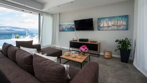 Ein modernes Wohnzimmer mit großem Fenster mit Blick auf ein Gewässer, einem Flachbildfernseher, zwei Gemälden, einem Couchtisch mit dekorativen Accessoires und einer Topfpflanze.