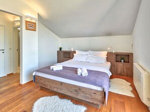 A bedroom with a wooden bed frame, white bedding, and a fluffy rug. There are two bedside tables with lamps and a small plant on one.