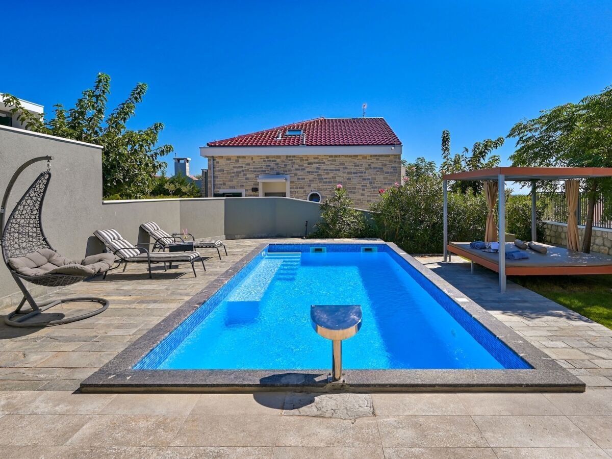 A rectangular swimming pool with a clear blue interior, surrounded by stone tiles. There are lounge chairs and a pergola with a roof on the right side.