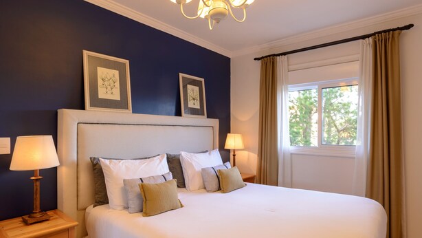 Una cama con sábanas blancas y varios almohadones, flanqueada por mesitas de noche con lámparas, en una habitación con paredes azul oscuro y un gran ventanal con cortinas.