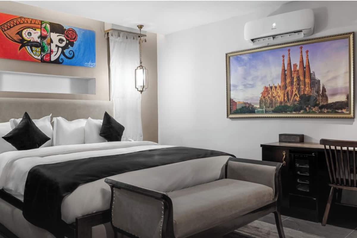 A modern hotel room with a large bed, a desk, a chair, a painting of a cathedral, and a colorful artwork above the bed.