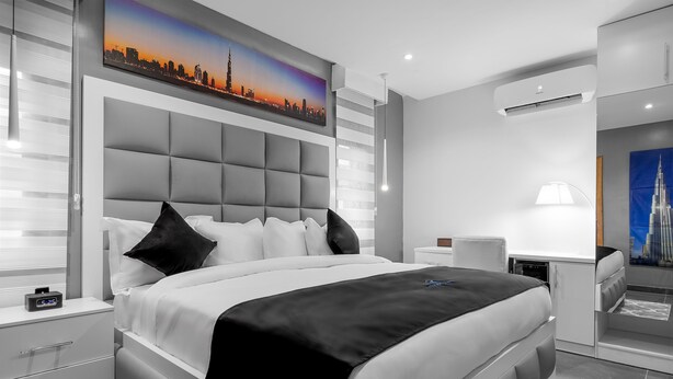 A modern bedroom with a large bed, a nightstand, a desk, and a chair. There is a cityscape painting on the wall and an air conditioning unit on the ceiling. - Villa Monument Hotel (Lagos)