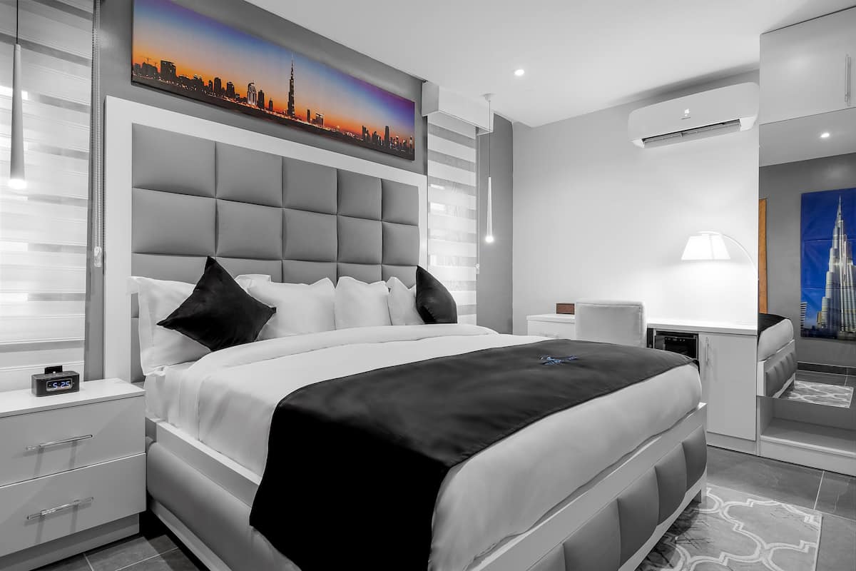 A modern bedroom with a large bed, a nightstand, a desk, and a chair. There is a cityscape painting on the wall and an air conditioning unit on the ceiling.