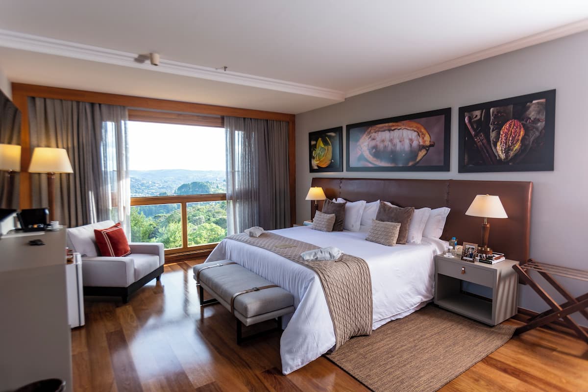 A hotel room with a large bed, a desk, a chair, a bench, and a view of the outdoors.