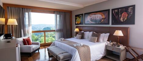 A hotel room with a large bed, a desk, a chair, a bench, and a view of the outdoors.
