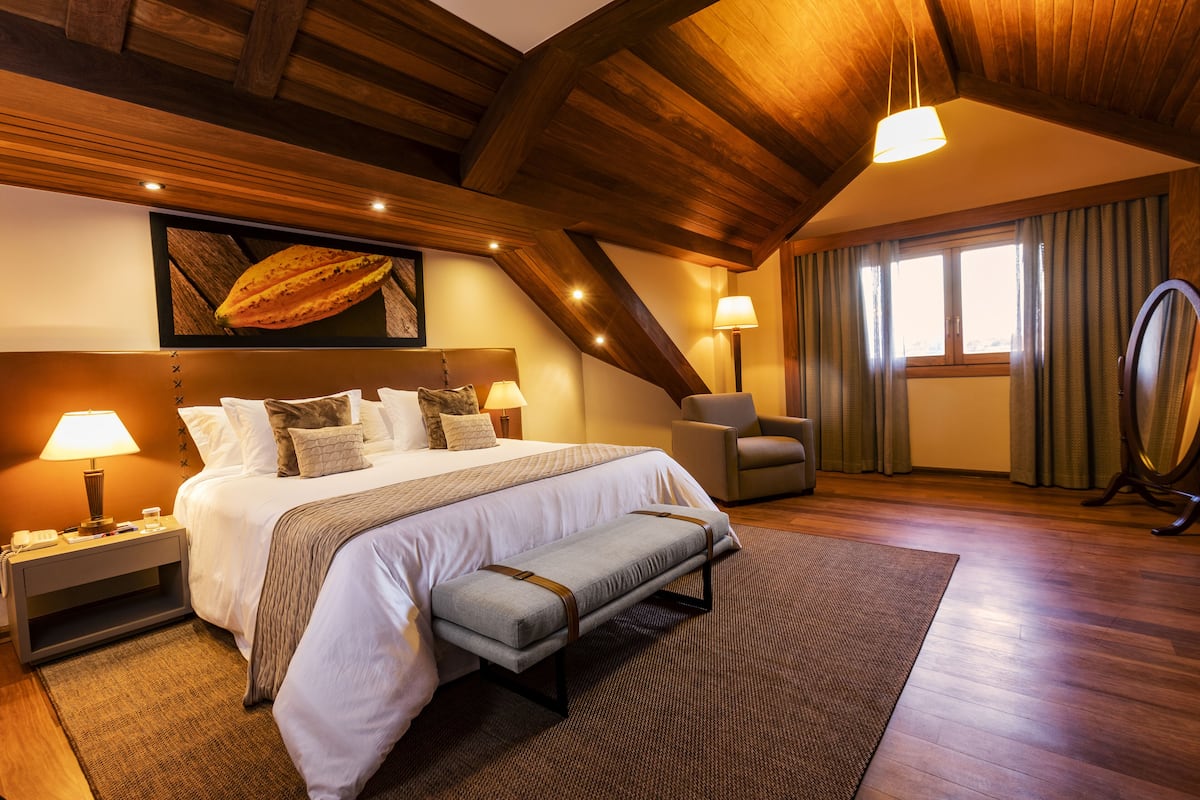 A spacious bedroom with a large bed, bedside lamps, a bench, a chair, and a large window with curtains.