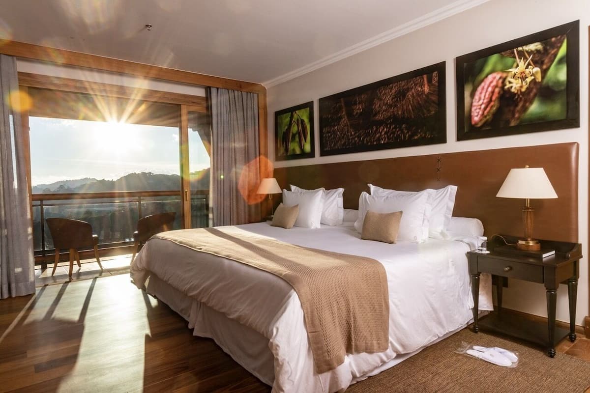 A hotel room with a large bed, two bedside tables, a view of the mountains, and artwork on the walls.
