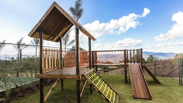 Uma estrutura de madeira para brincar, com escorrega e rede de escalada, cercada por uma grade e gramado, com vista para colinas ao fundo.