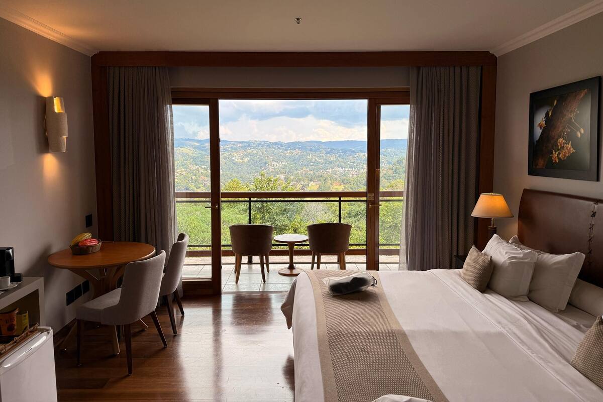 A hotel room with a large bed, a desk, chairs, and a balcony with a scenic view.