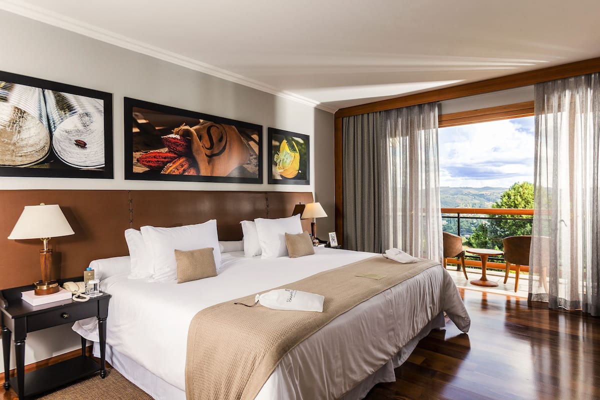 A hotel room with a large bed, a nightstand, a lamp, a phone, a framed picture, and a balcony with a view.