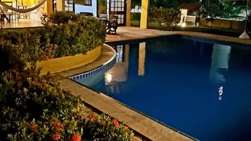 Uma área de piscina à noite, com um prédio bem iluminado, uma rede e um grande elemento aquático.