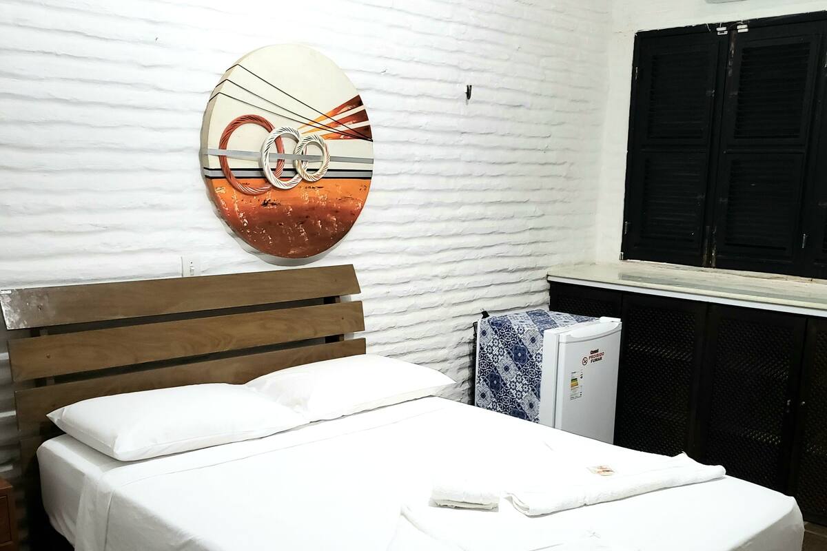A neatly made bed with white linens and towels, a wooden headboard, a wall-mounted air conditioner, and a window with black shutters.