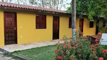 Um edifício amarelo com várias portas e janelas, rodeado por um espaço pavimentado e um pequeno jardim com flores vermelhas.