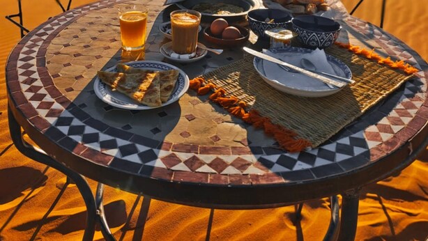 A dining table with a traditional Moroccan spread, including bread, tea, and a teapot, set against a backdrop of a desert landscape with sand dunes and a clear sky. - dune merzouga camp (Rissani)