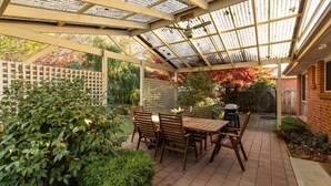 Un patio couvert, agrémenté d’une table et de chaises, entouré de verdure et bordé d’un mur en briques.
