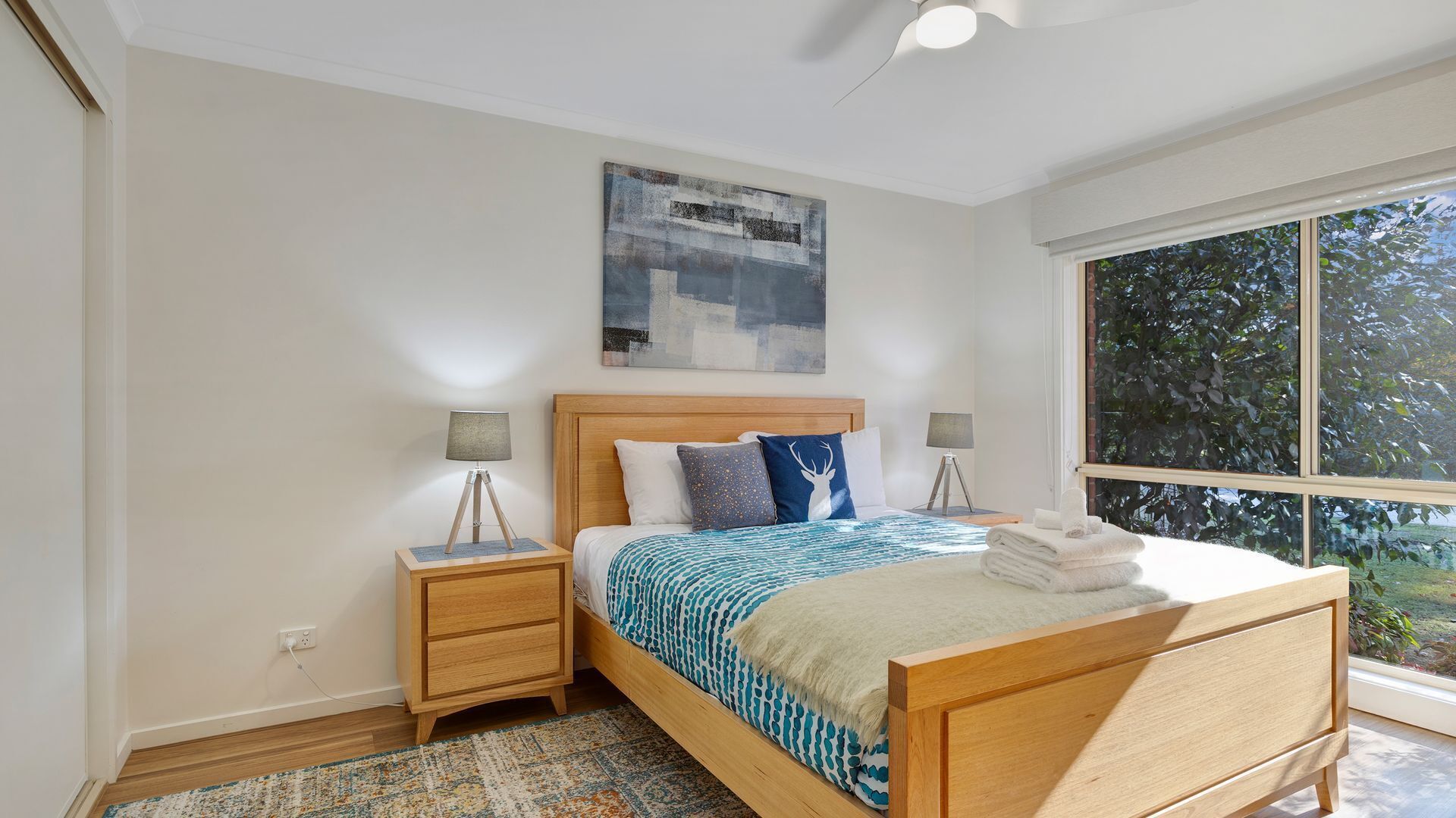 A bedroom with a wooden bed, two bedside tables, a floor lamp, a large window, and a painting on the wall.