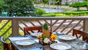 Une table dressée pour un repas, agrémentée d’un ananas en guise de centre de table, entourée de chaises et d’assiettes, donnant sur un espace aménagé avec des palmiers et un bâtiment en arrière-plan.